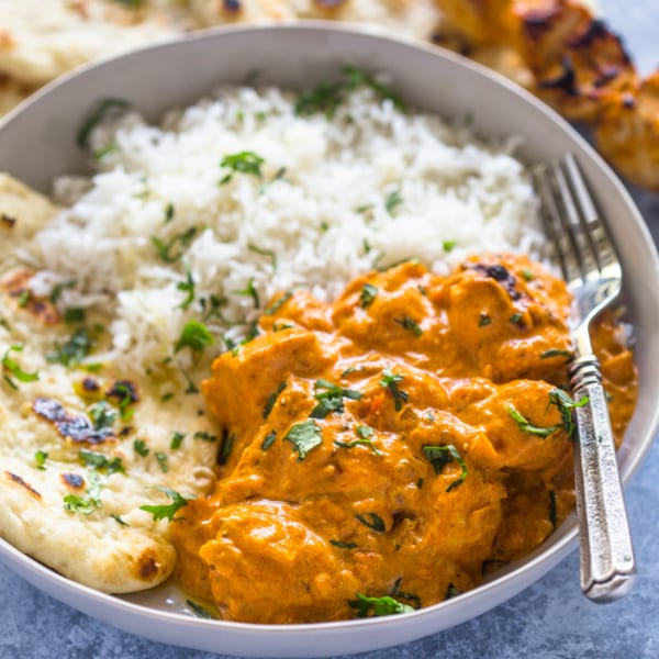 A bowl of butter chicken with white rice, naan bread, and a fork.