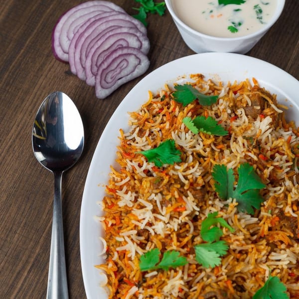 A plate of biryani with cilantro, sliced red onions, and a bowl of raita on a wooden table.