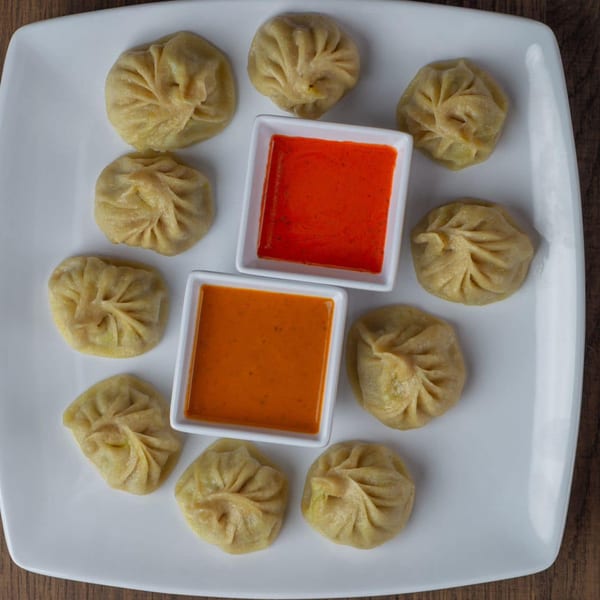 A white square plate holds ten steamed dumplings surrounding two small bowls of red and orange dipping sauces.