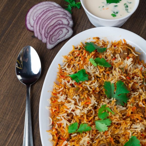 Biryani with cilantro, sliced red onions, and a side of raita on a wooden table.