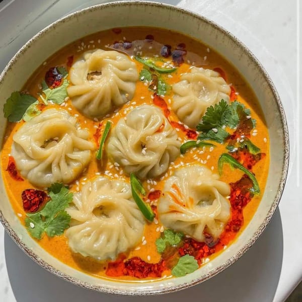 A bowl of momos in a vibrant orange sauce, garnished with cilantro, chili oil, and sesame seeds.