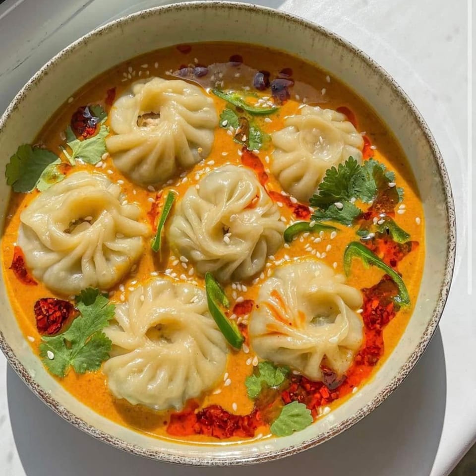 A bowl of momos in a vibrant orange sauce, garnished with cilantro, chili oil, and sesame seeds.