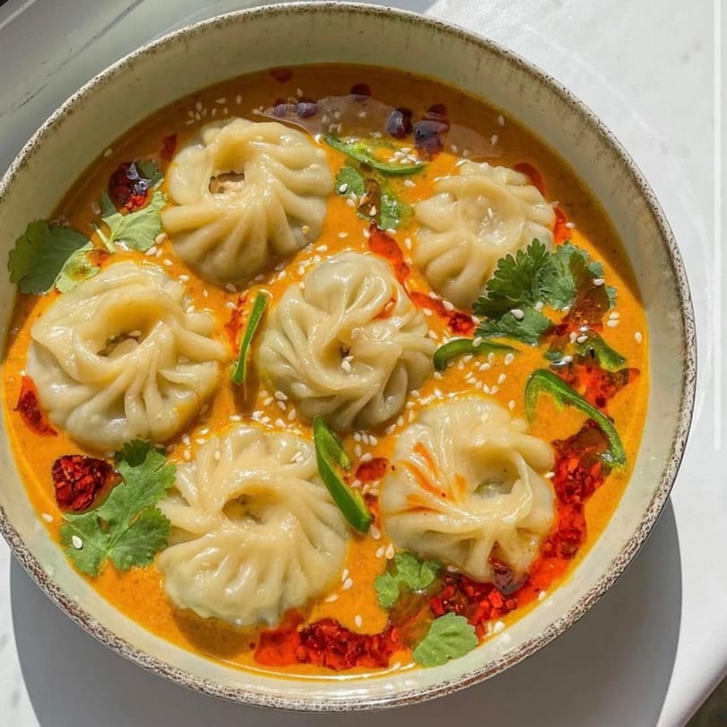 A bowl of momos in a vibrant orange sauce, garnished with cilantro, chili oil, and sesame seeds.